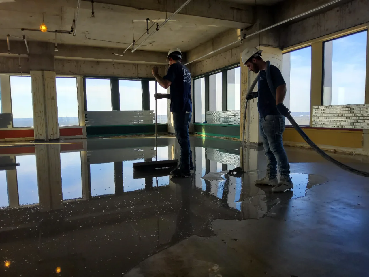 Two installers spreading self-leveling underlayment with gauge rakes and a smoother in front of the windows